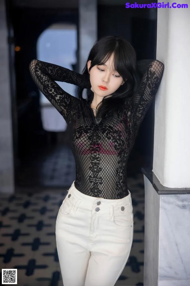 A woman in a black top and white pants leaning against a wall.