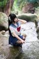 A woman in a school uniform sitting on a rock by a stream.