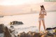 A woman in a white bikini standing on rocks by the water.